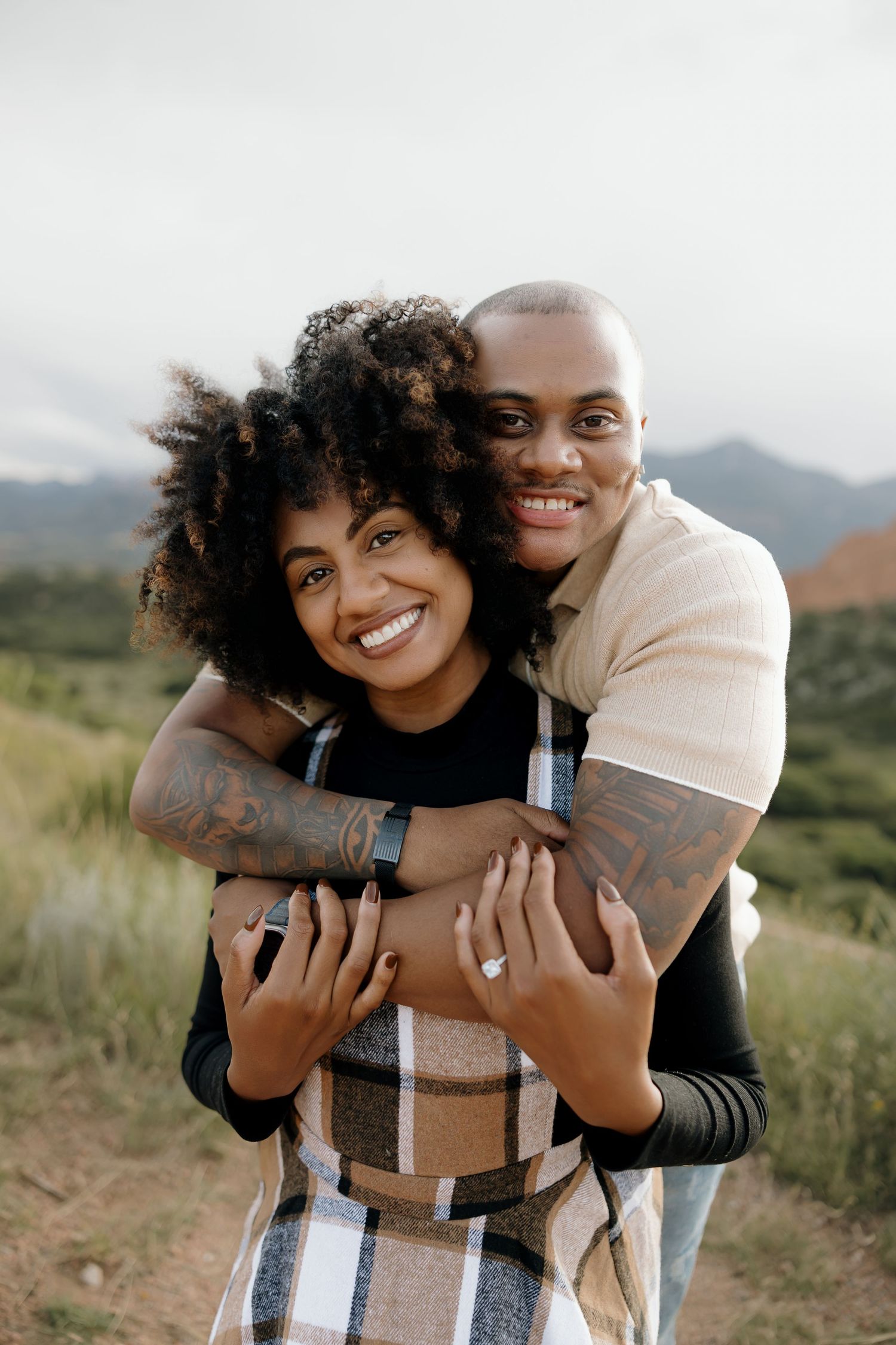 A couple embraces and laughs together in an outdoor setting with mountains visible in the background.