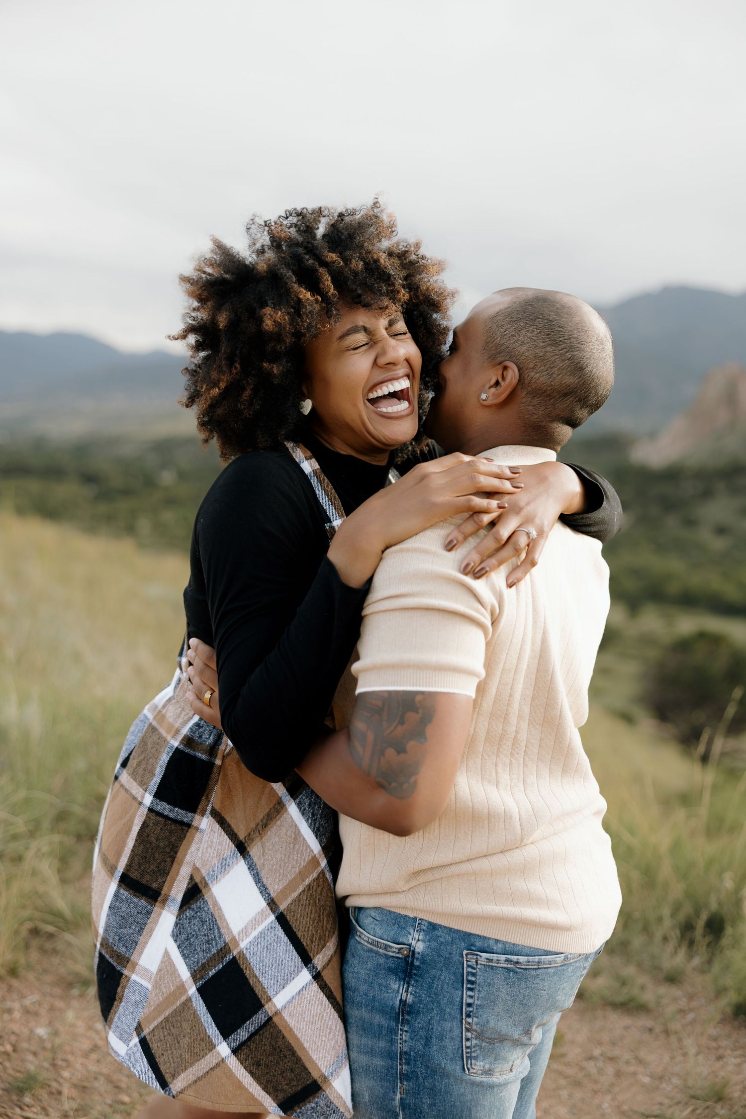 A joyful couple embraces and laughs together while standing in a grassy field with mountains in the background.