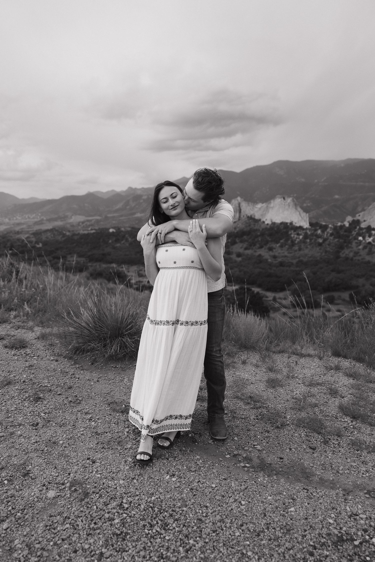 A romantic black and white photo shows a couple embracing on a mountain overlook with scenic hills in the background.