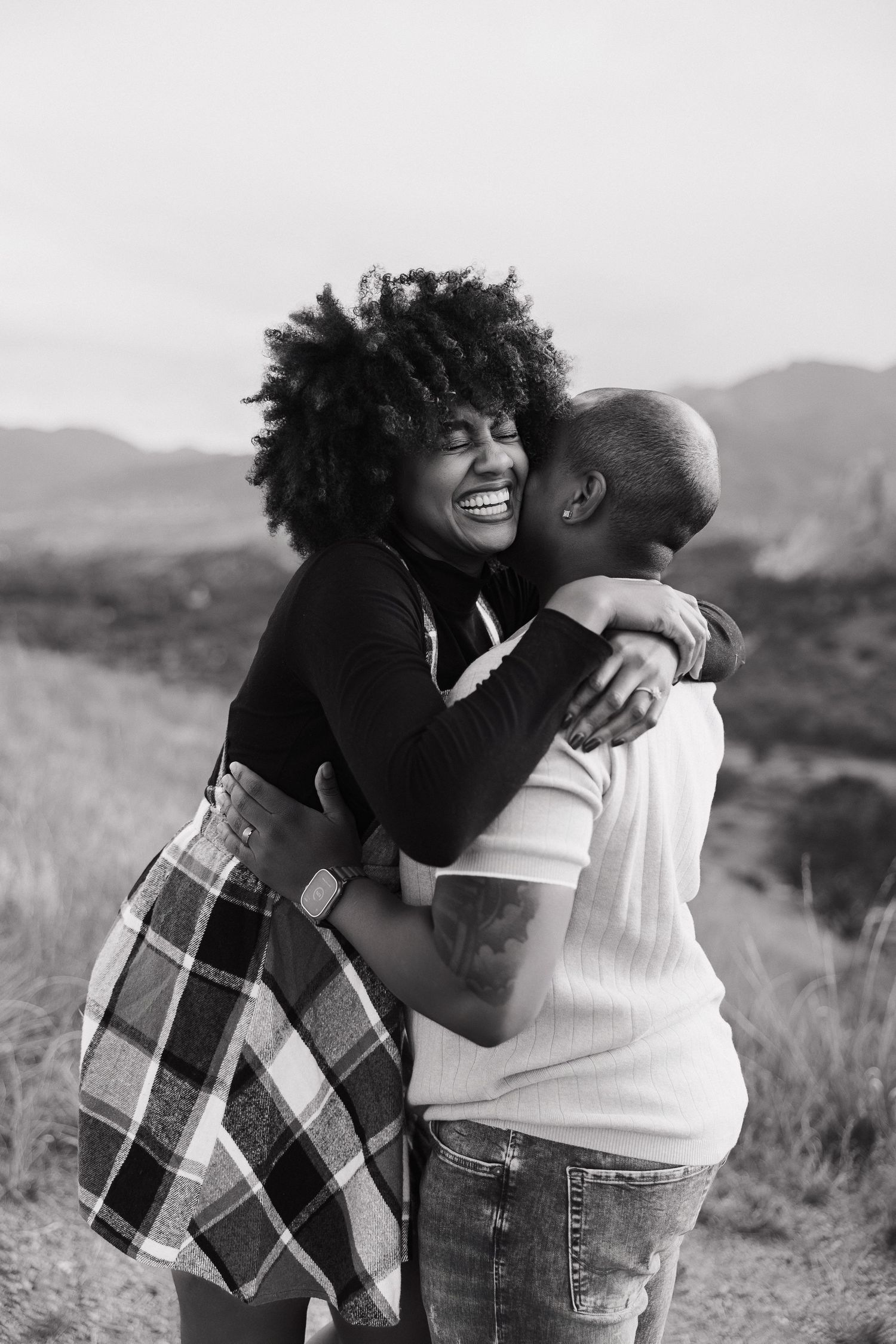 A loving couple embraces and laughs together in a scenic outdoor setting with mountains in the background.