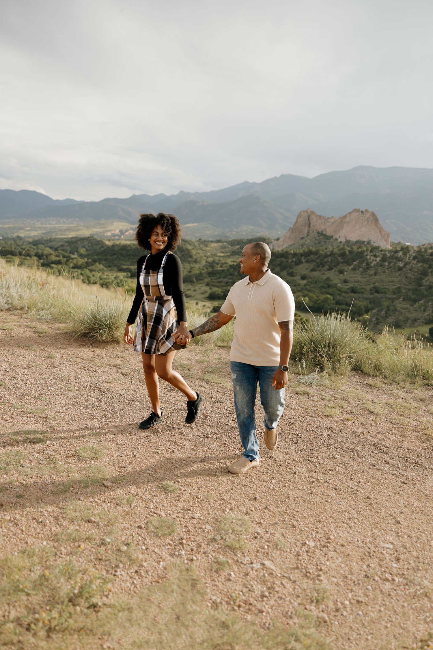 Two people walk along a desert trail with mountains in the background under cloudy skies.