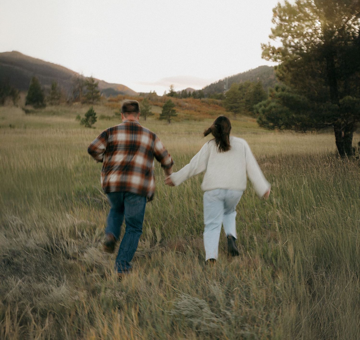 Couple walking hand in hand through a golden meadow at sunset with mountains in the background.