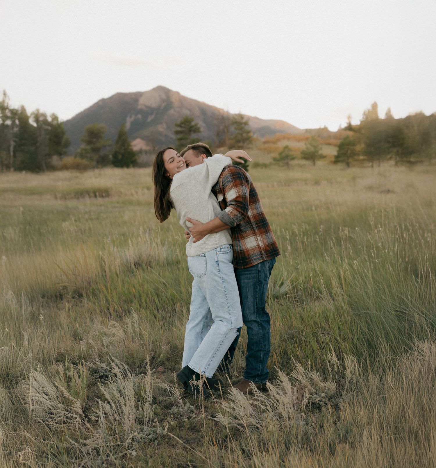 A couple embraces in a grassy field with mountains and trees in the background during golden hour.