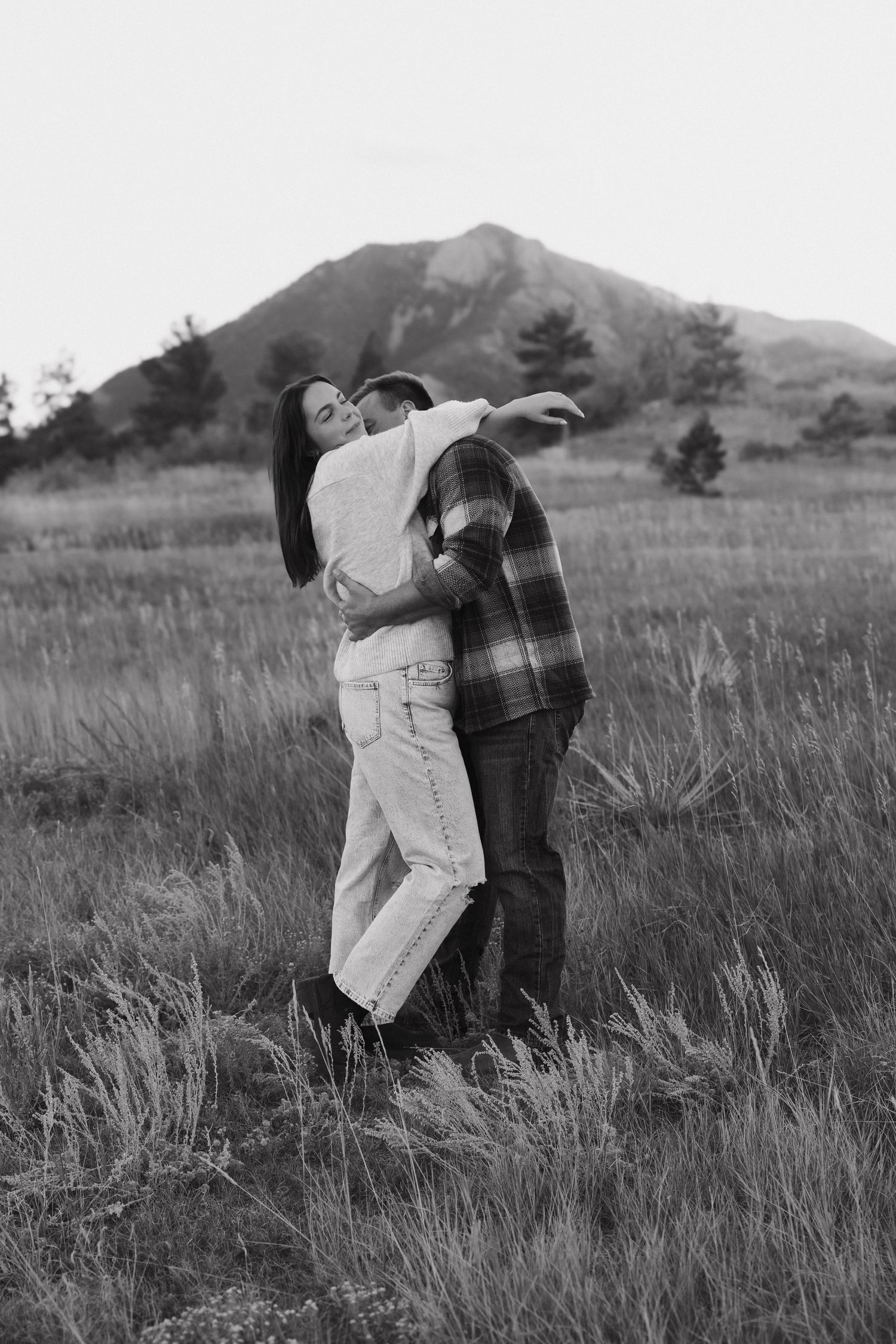 A romantic black and white photo series showing a couple embracing in a grassy field with mountains in the background.