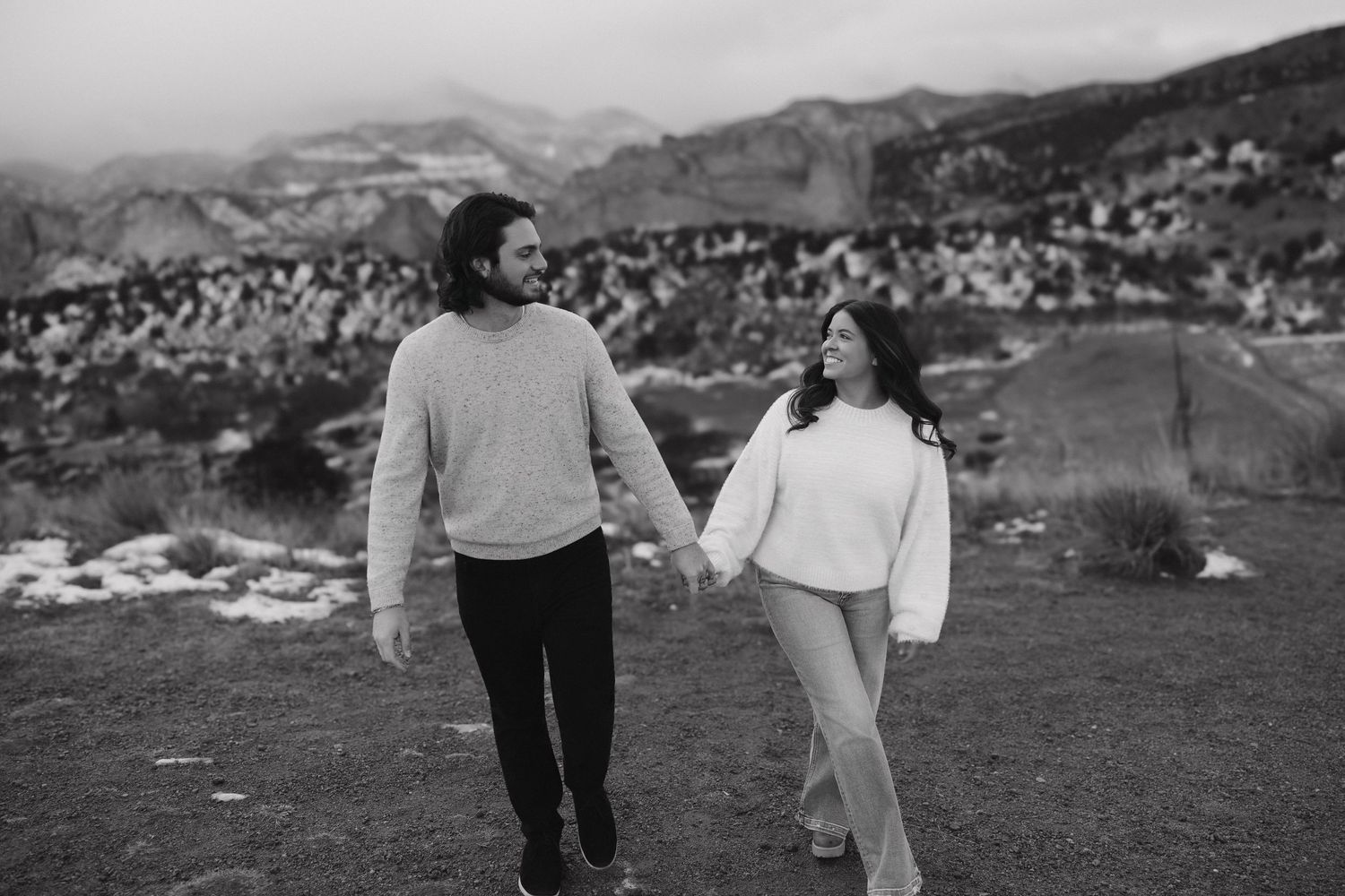 A couple walks hand in hand through a scenic mountain landscape on a cloudy day.