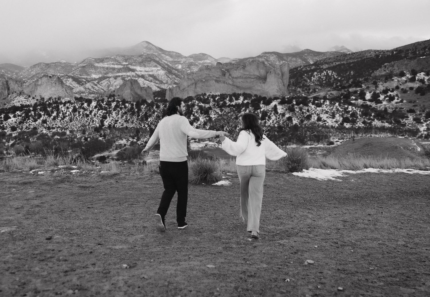 Two people walk along a rocky mountain path with dramatic mountain peaks rising in the background.