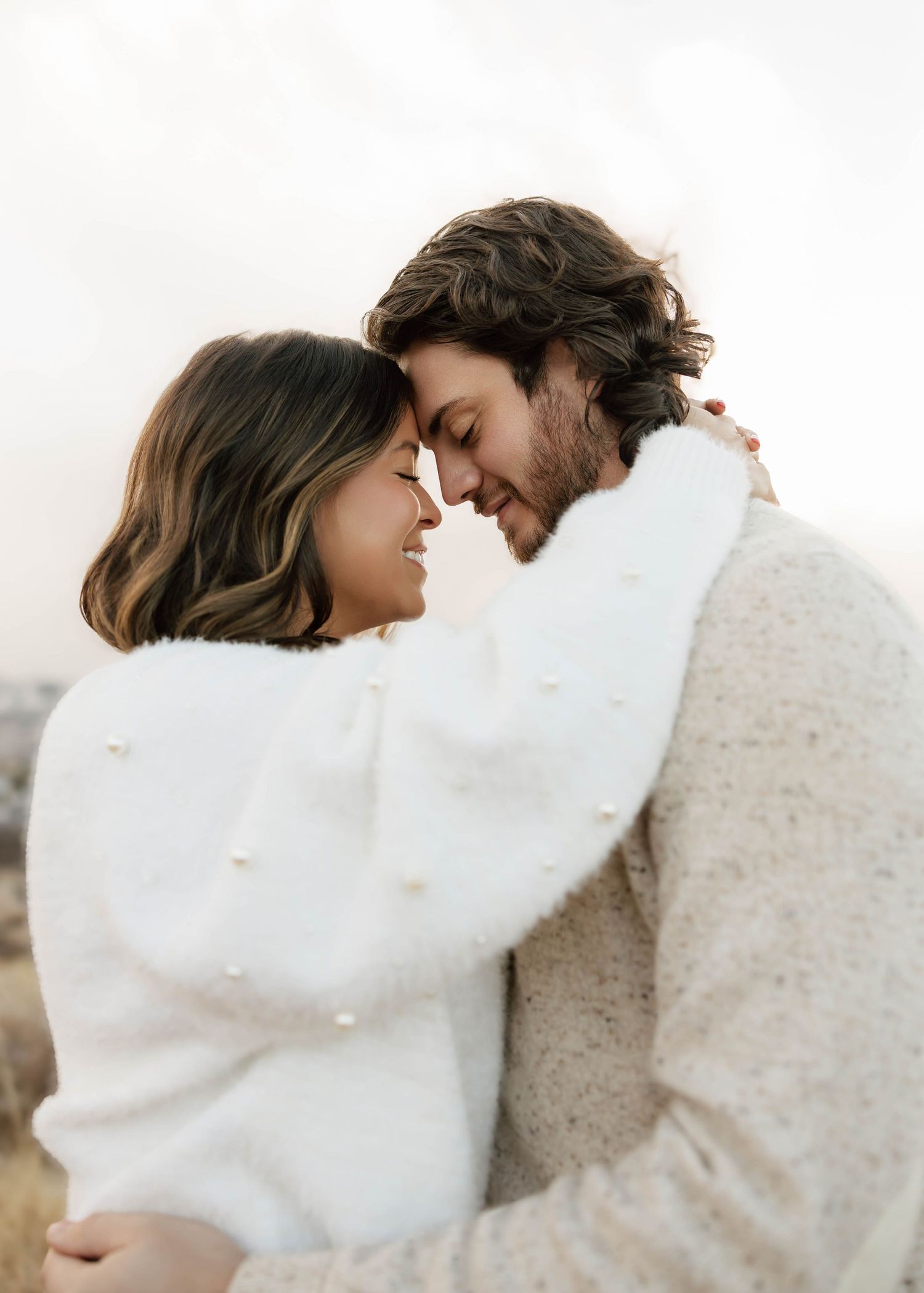 A couple in white and beige outfits embrace intimately against a bright background in a romantic photoshoot.