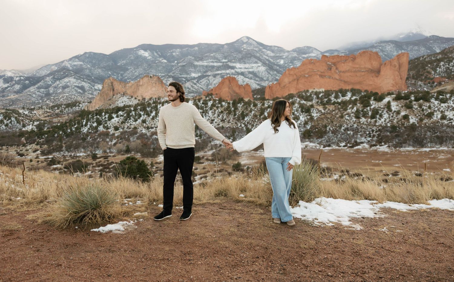 Two people hold hands in front of snowy Garden of the Gods rock formations in Colorado Springs during winter.