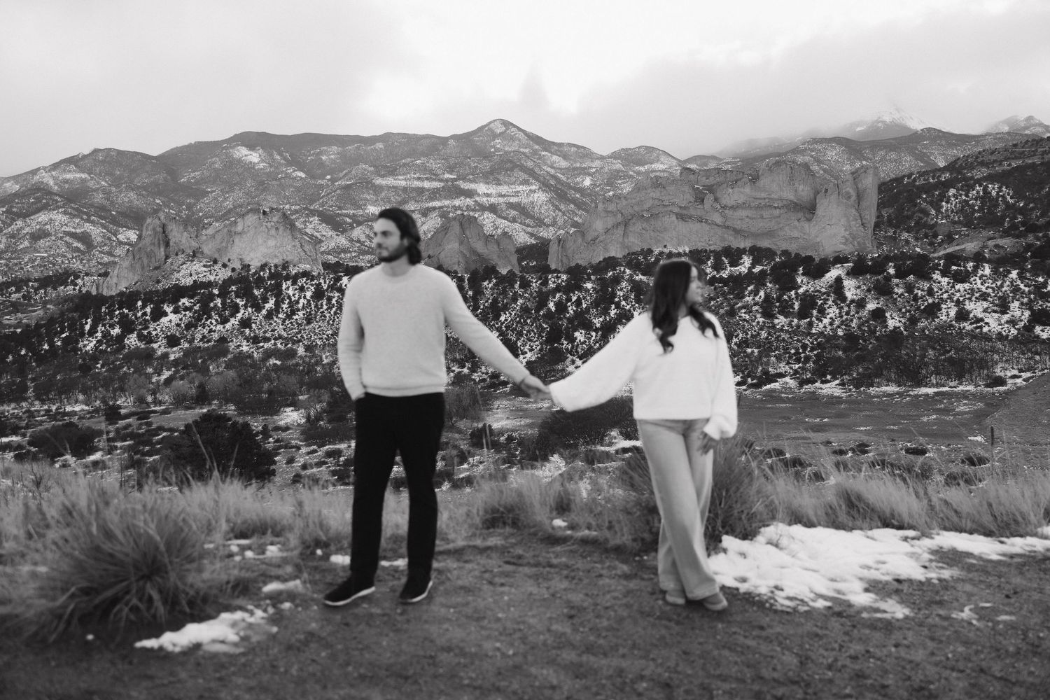 A couple holds hands while standing on a snowy mountain landscape in black and white photography.