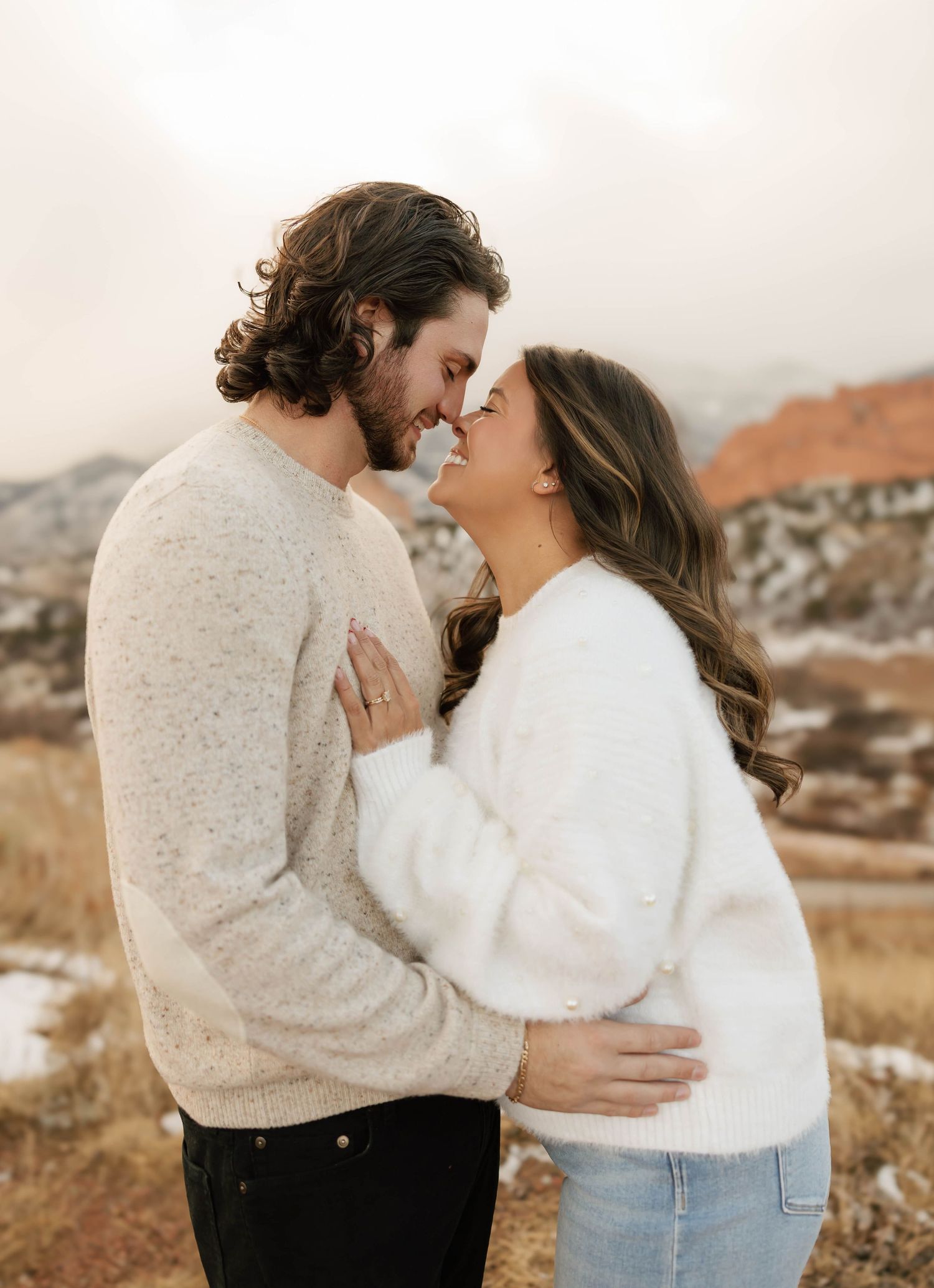 A romantic couple in cozy sweaters share an intimate moment against a scenic mountain backdrop in warm autumn light.