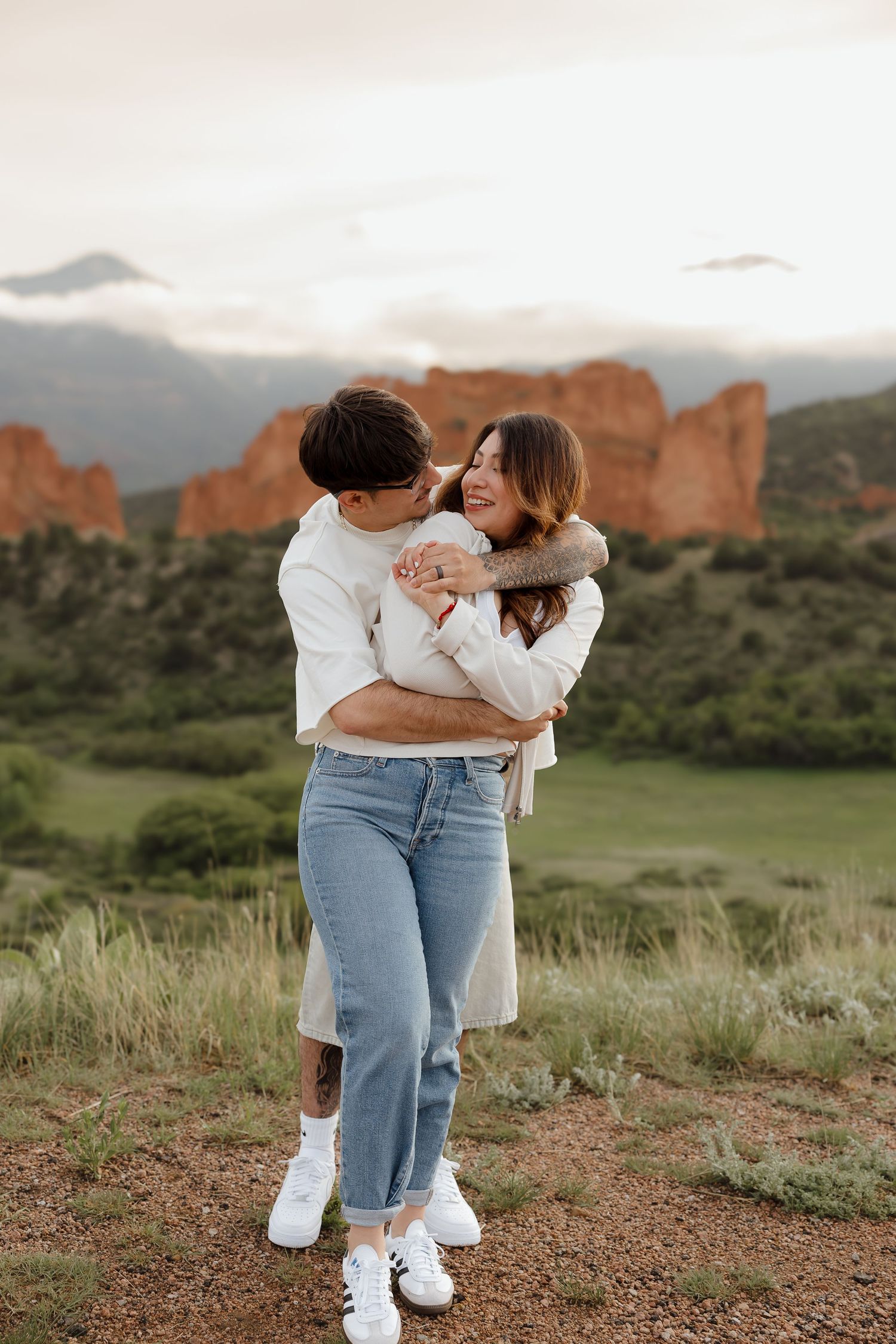 Couple in white and denim embracing on a hill with red rock formations and mountains in the background at sunset.
