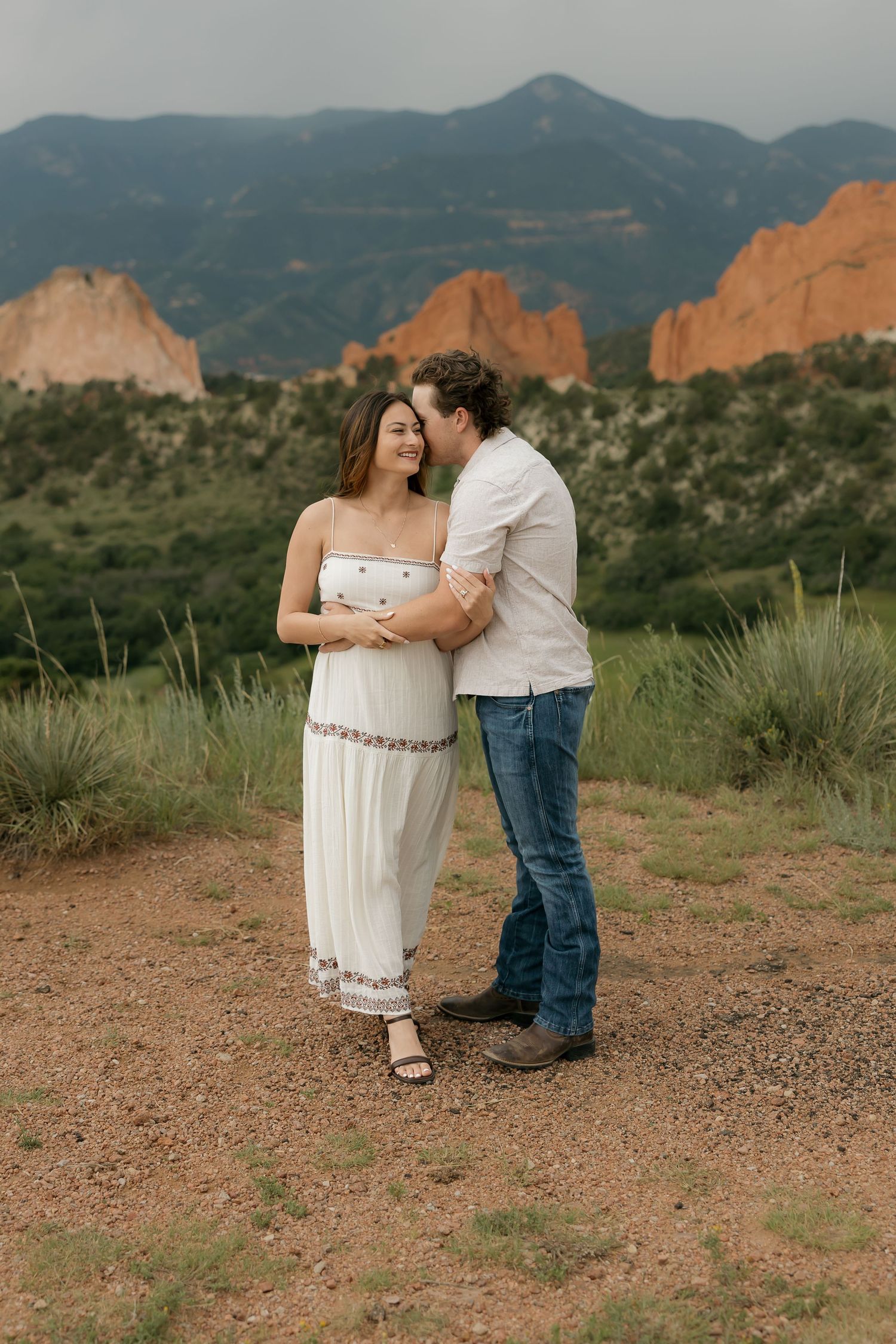 A couple embraces at Garden of the Gods in Colorado Springs with red rock formations and mountains in the background.