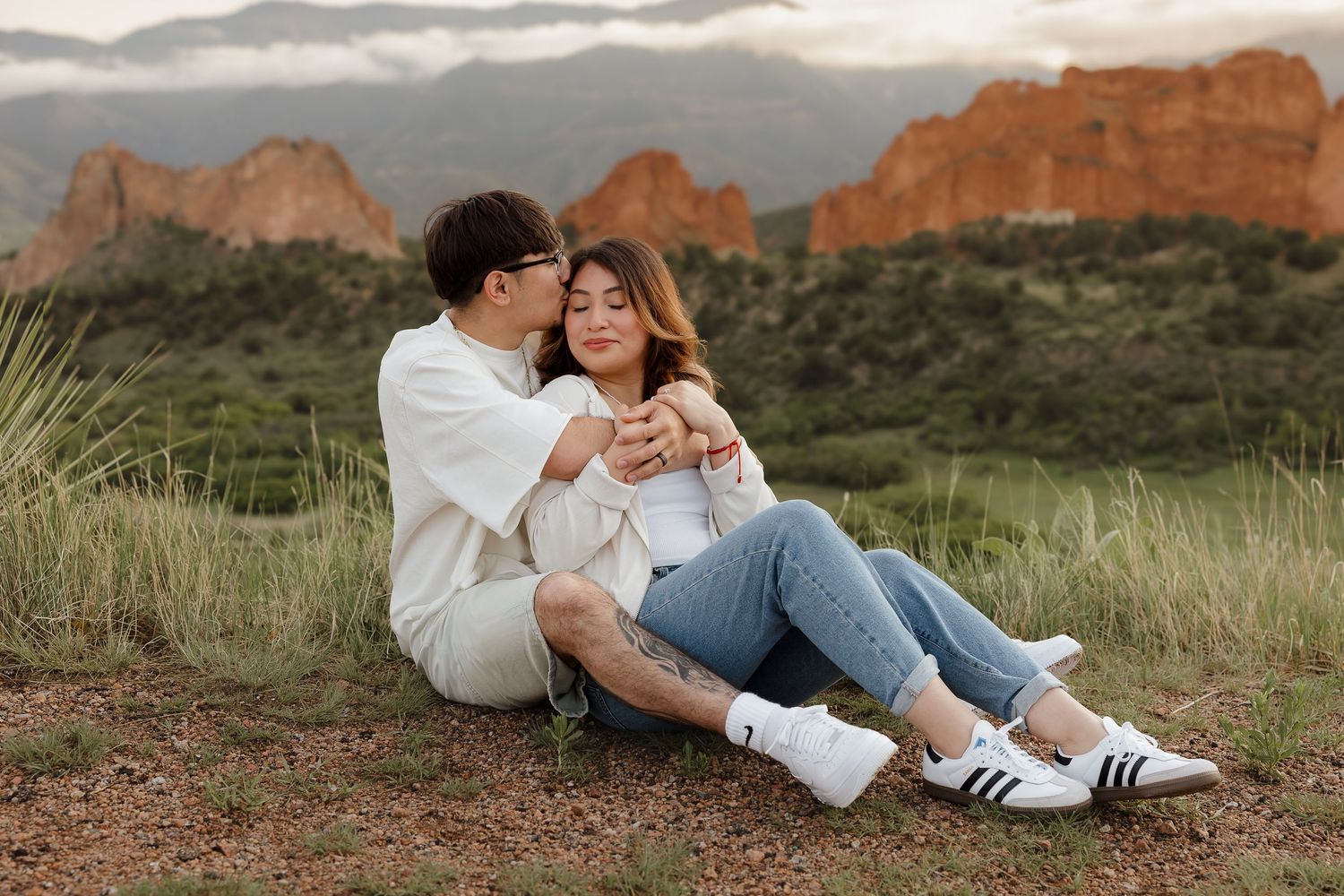 A couple sits together on a grassy hillside with majestic red rock formations and mountains in the background.