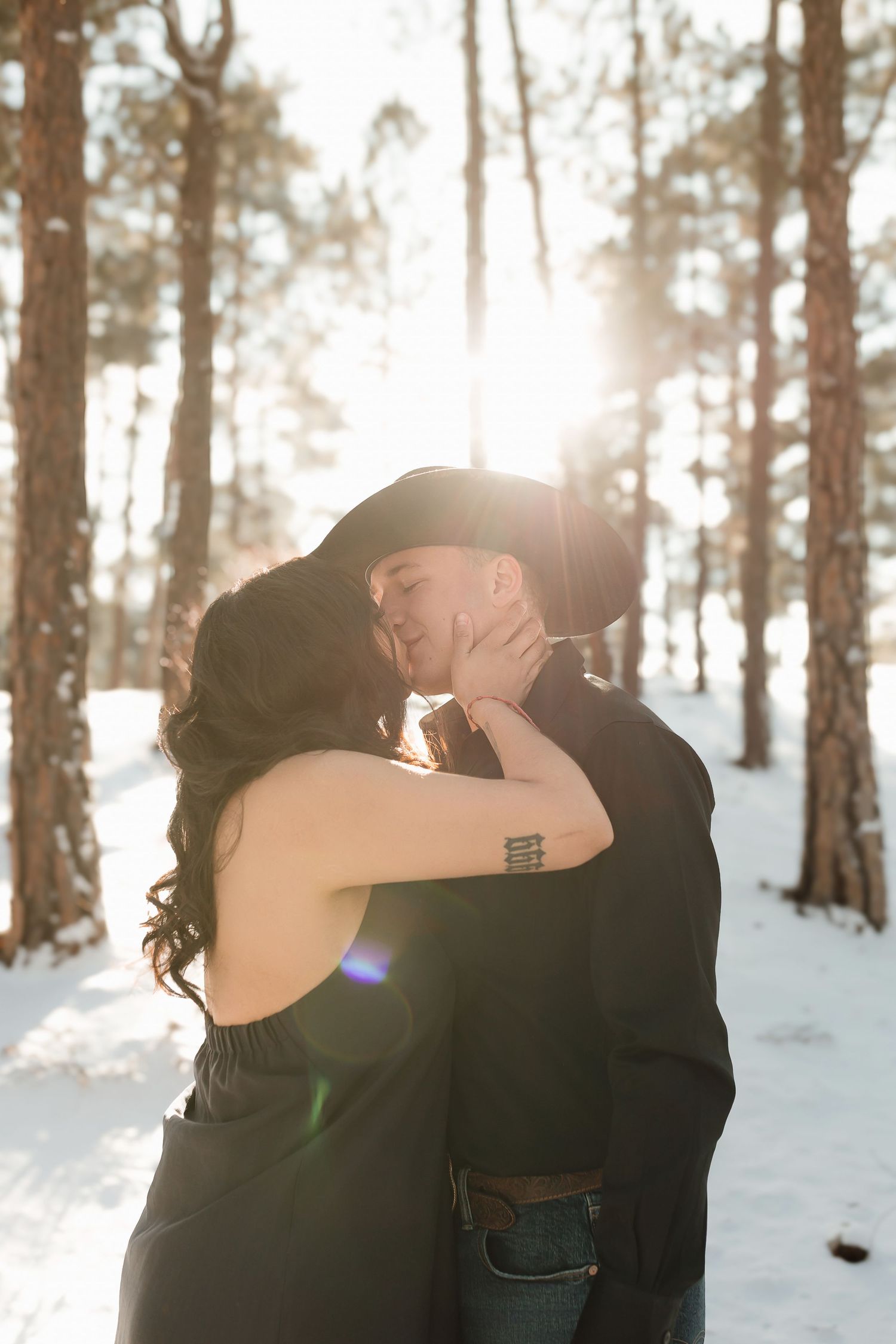 Romantic winter engagement photo silhouetted against bright sunlight streaming through a snowy forest.