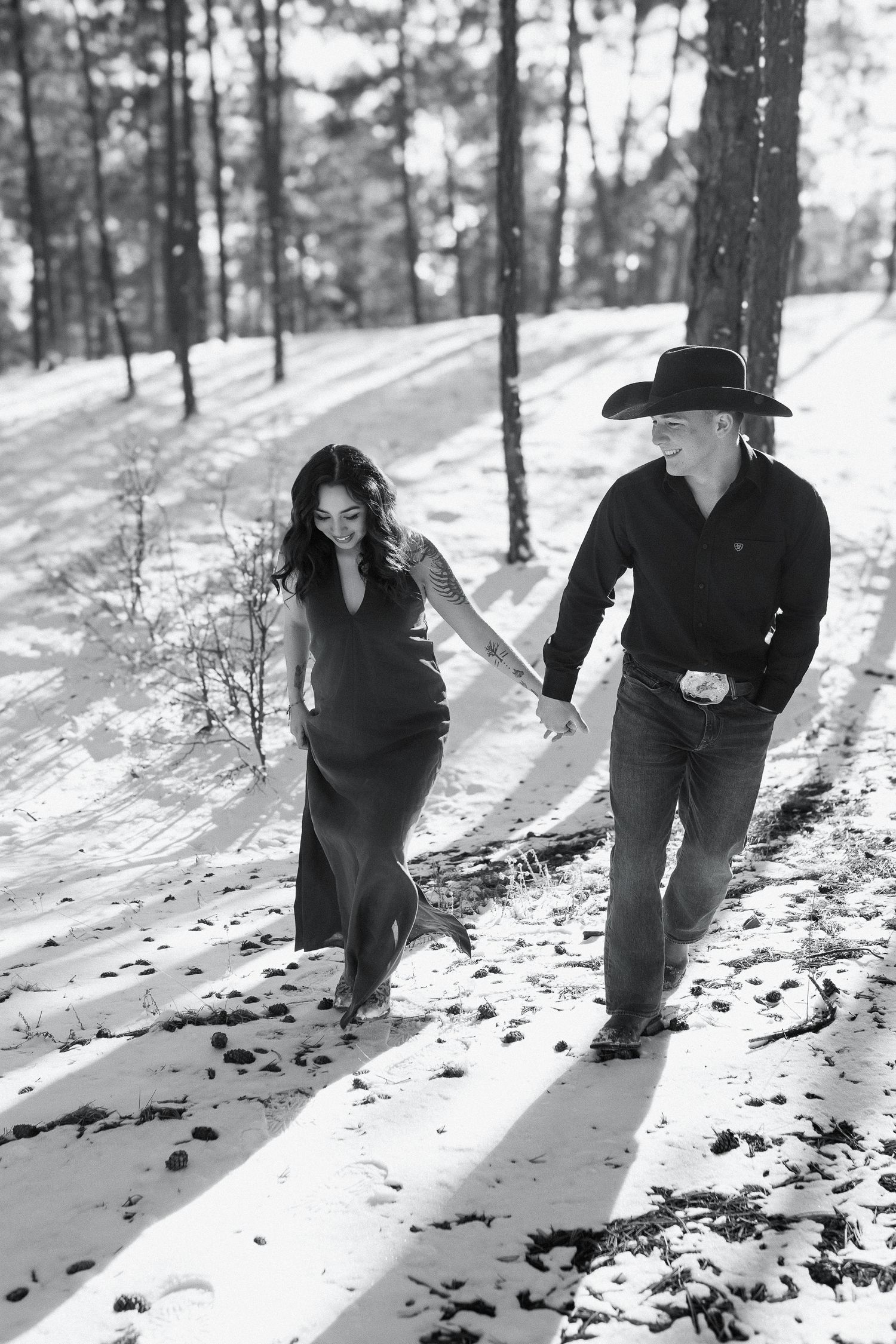A couple in western style attire walk together holding hands through a snowy forest path on a winter day.