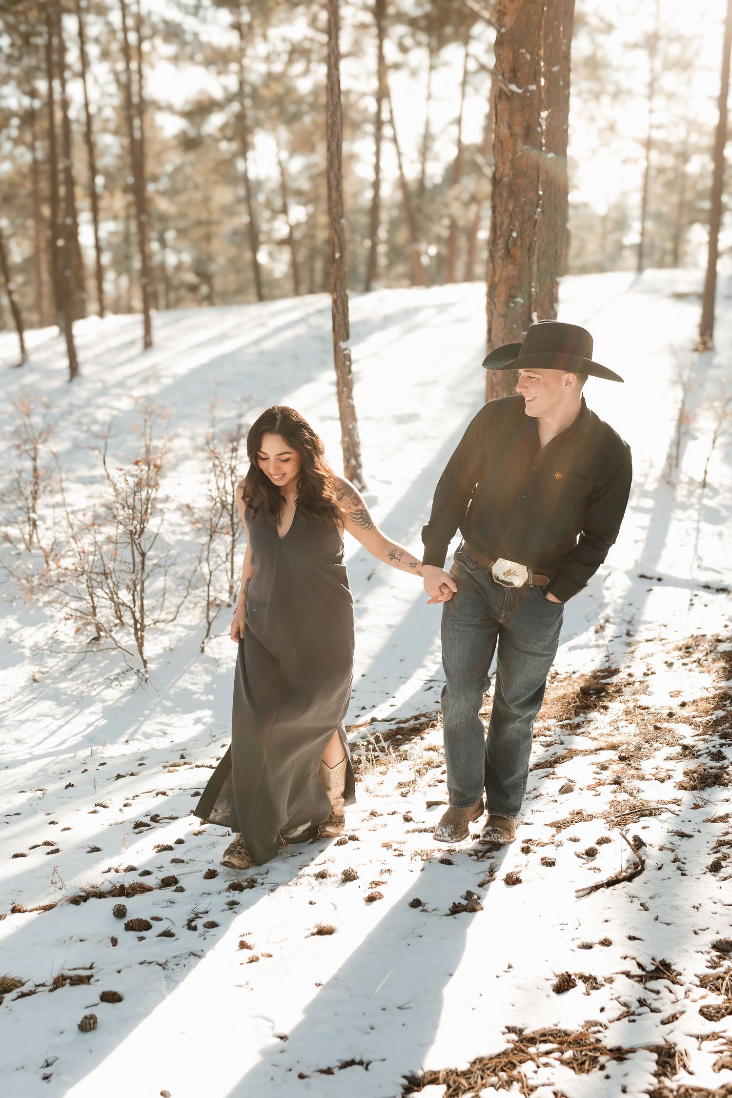 A couple stands in a snowy winter forest with sunlight streaming through tall trees creating long shadows on white ground.