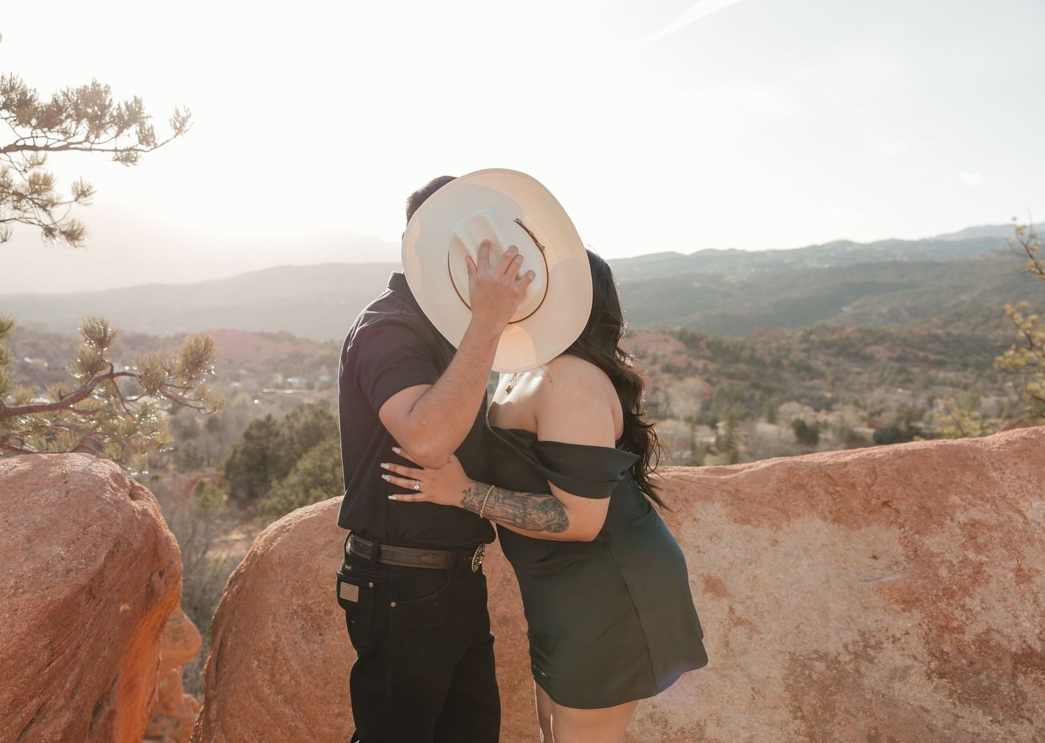 Two people share a romantic sunset moment while overlooking mountain vistas from a rustic red rock overlook point.