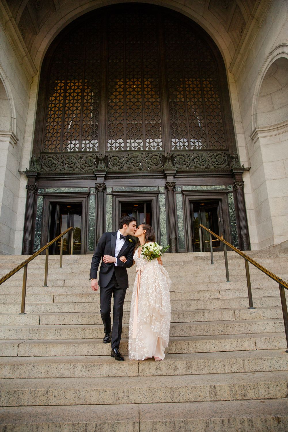 A wedding couple poses on the ornate stone steps of a building with decorative iron doors.