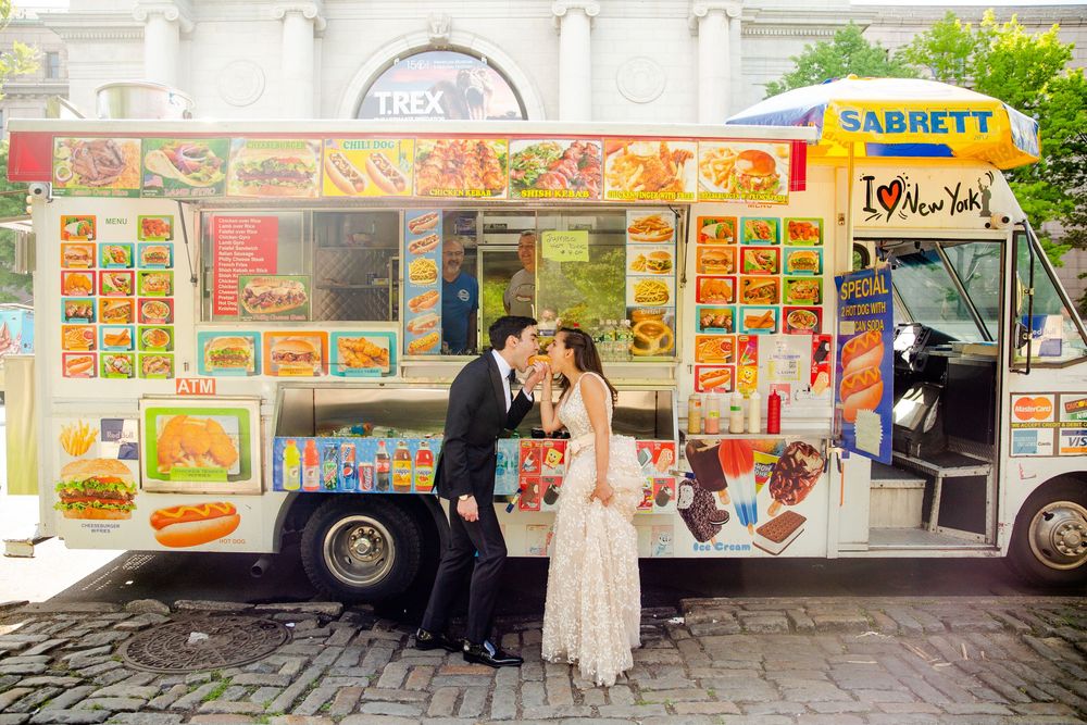 A couple stands in formal attire next to a vibrant food truck serving various menu items on a city street.