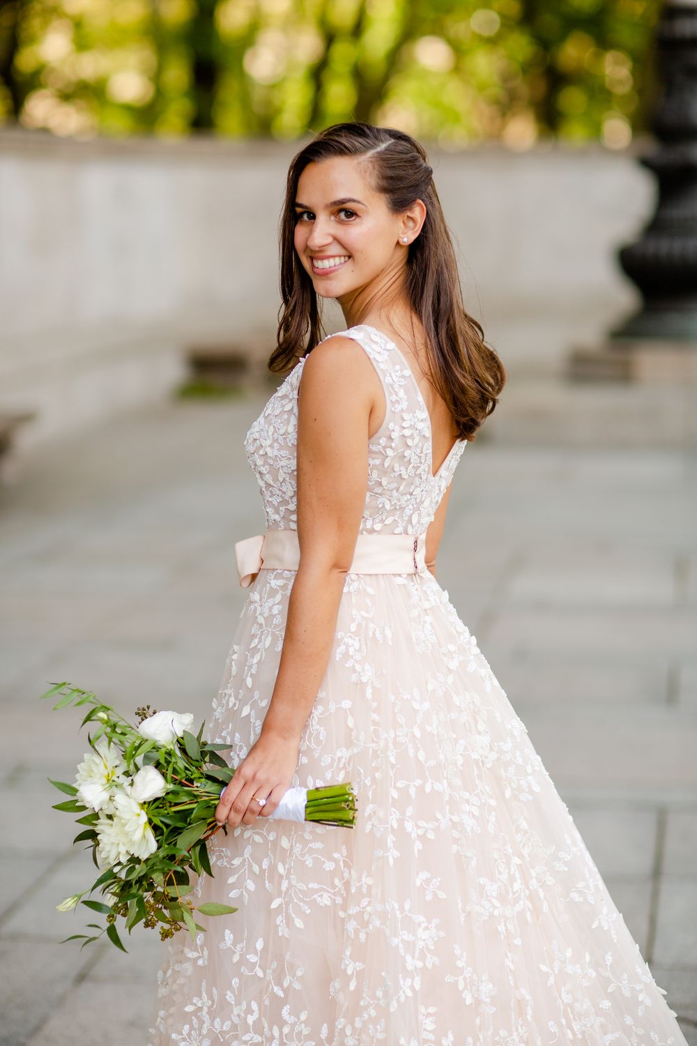 An elegant bride in a delicate lace wedding dress holds a white floral bouquet against a soft bokeh background.