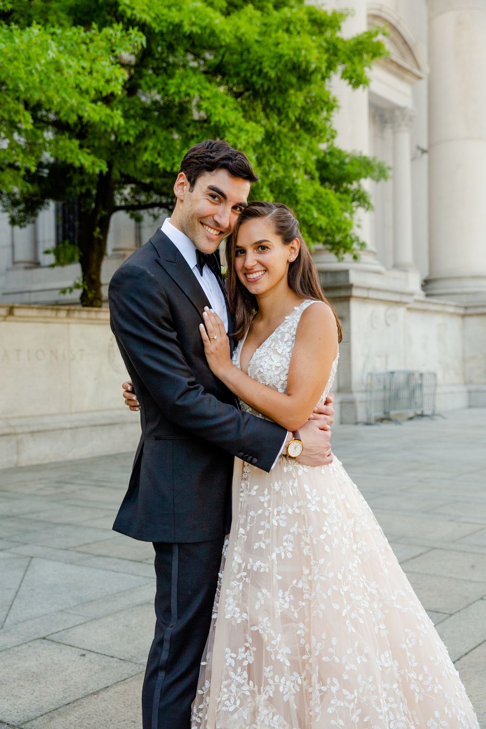 A romantic wedding portrait in front of a classic stone building with lush green trees.