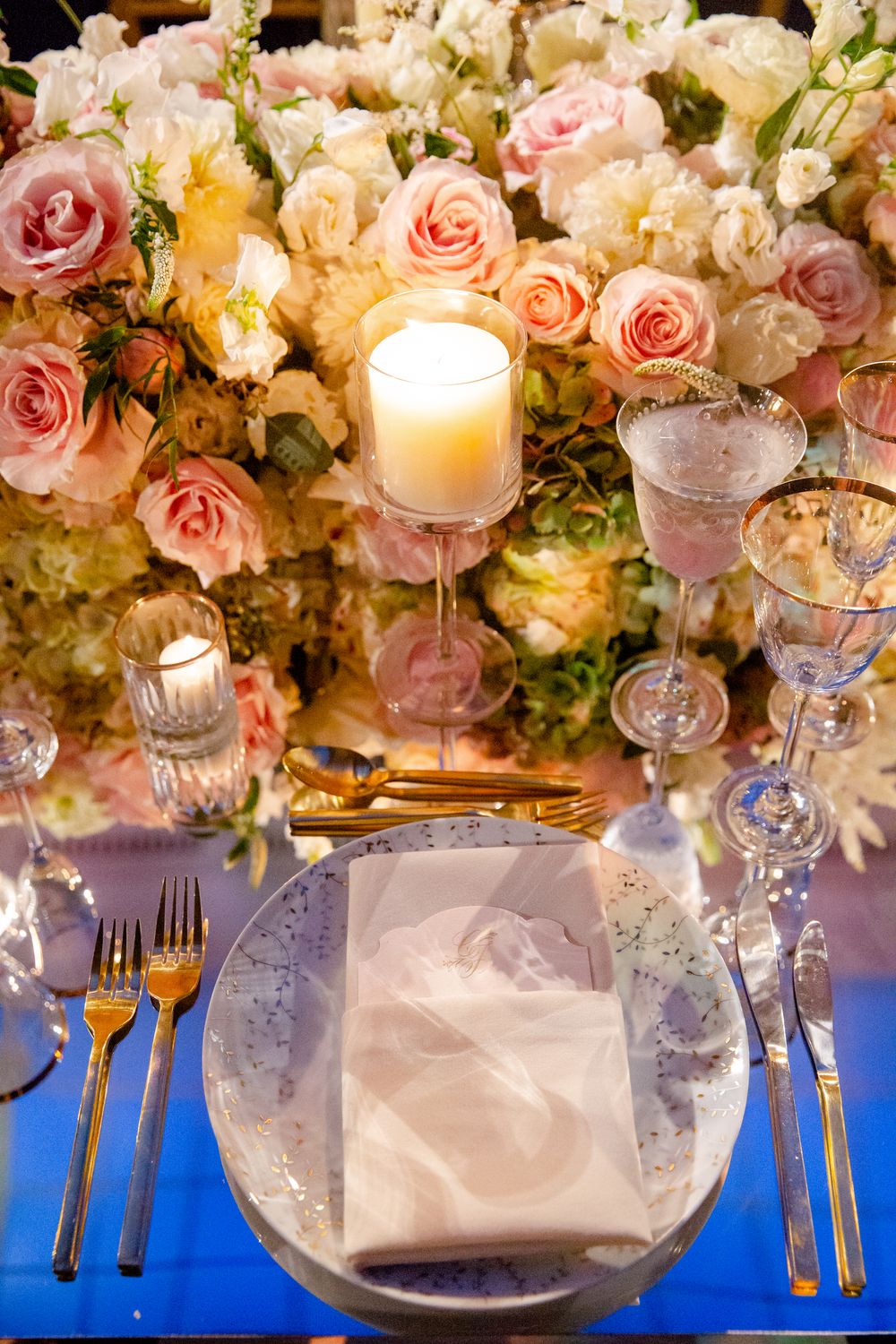Romantic table setting with pink roses, lit candles, and elegant place settings on blue tablecloth.