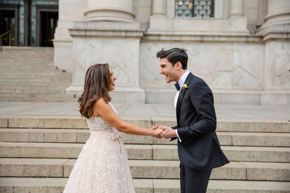 A couple in formal attire shares a joyful moment while dancing on stone steps outside a grand building.