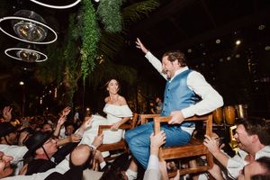Wedding reception crowd enthusiastically lifts people up during dance celebration at nighttime venue with greenery decorations.