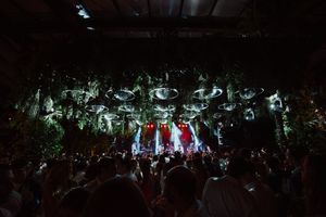 Concert performers stand on stage amid dramatic lighting effects and a large crowd at a live music performance.
