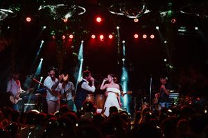 Performers on stage during a concert with dramatic blue lighting and red spotlights illuminating the dark venue.