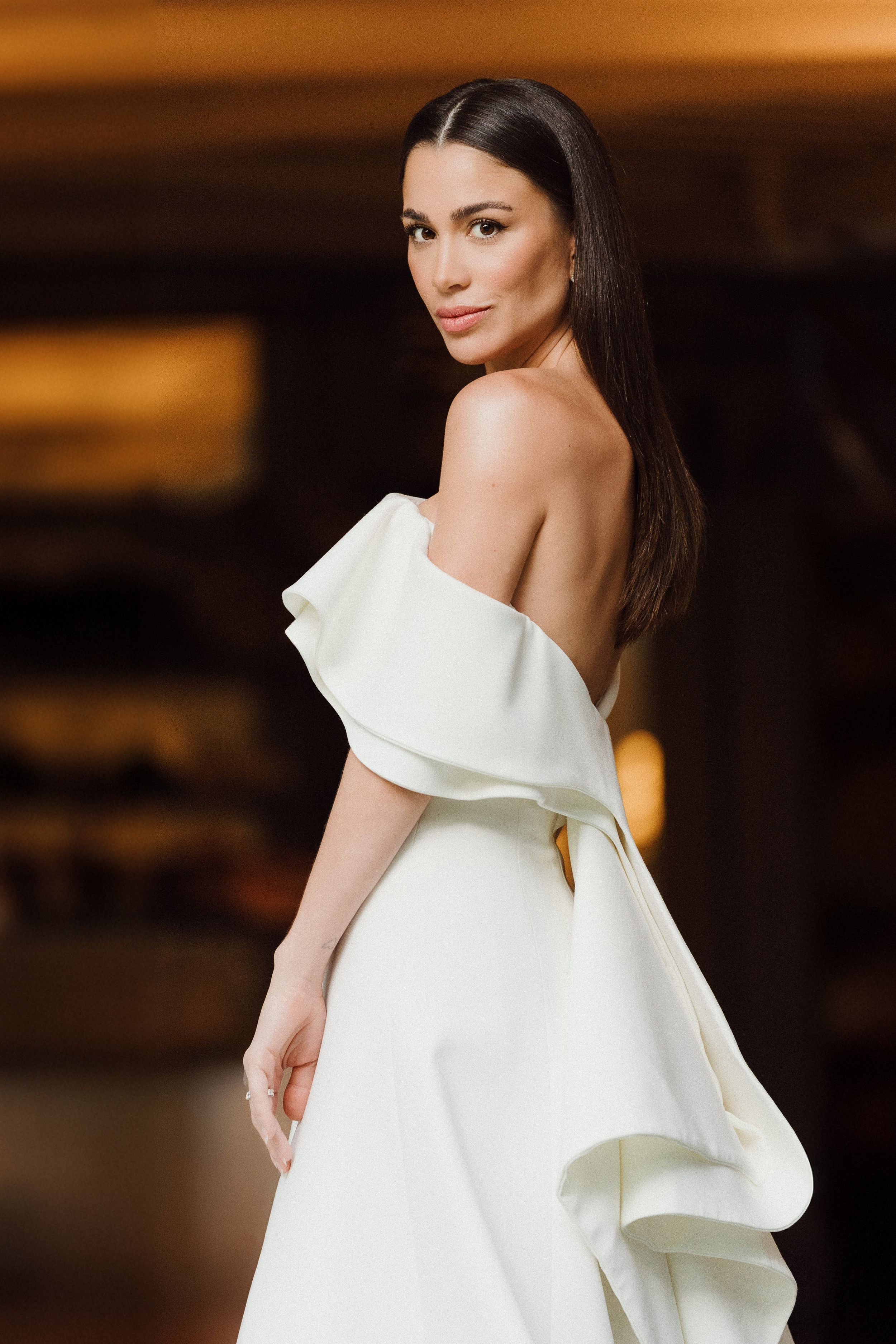 Model wearing elegant white asymmetrical evening gown with dramatic ruffled neckline detail against warm-toned background.