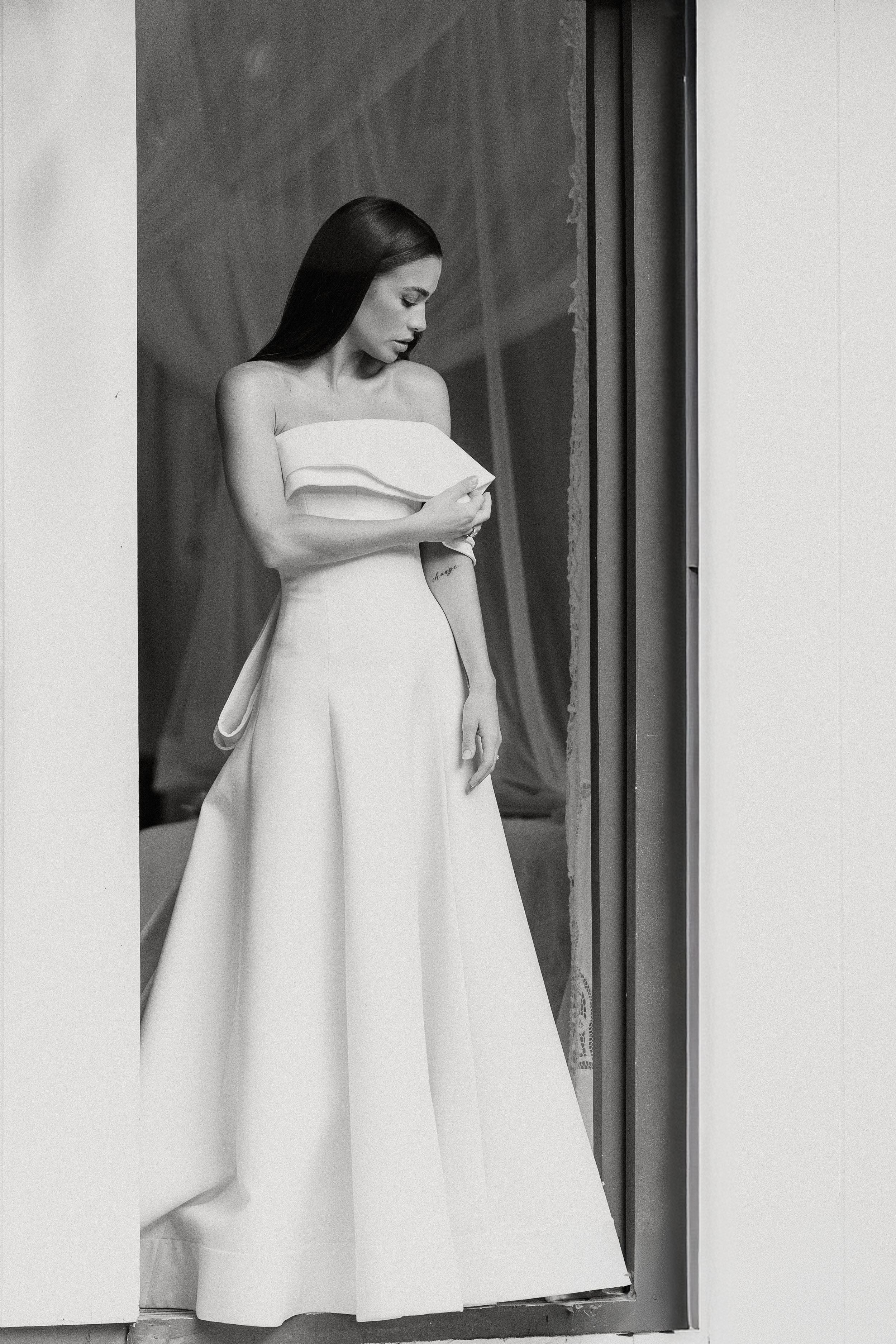 Classic black and white portrait series showing a bride in an elegant off-shoulder wedding dress looking through window light.