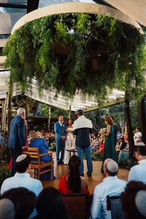Outdoor wedding ceremony under a lush green canopy with guests gathered around a wooden deck during daytime.
