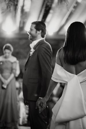 Black and white wedding photo capturing a couple holding hands during their ceremony with a bridesmaid in the background.