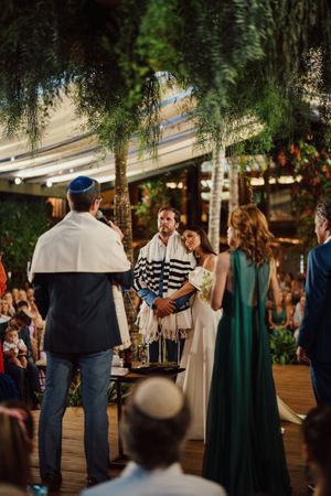 An outdoor Jewish wedding ceremony takes place under hanging greenery with guests gathered on a wooden deck at sunset.