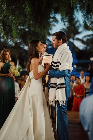 Elegant wedding ceremony outdoors under trees with bride in white gown and groom wearing traditional prayer shawl.