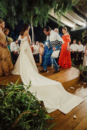 Wedding celebration on wooden dance floor under hanging greenery with elegant white gown and guests in festive attire.