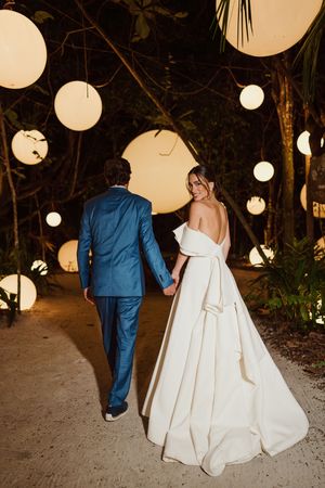 Couple walks down illuminated garden path with white lanterns overhead, bride in elegant gown and groom in blue suit.
