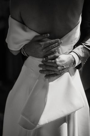 A close-up black and white image shows hands gently resting on someone wearing an elegant white dress.
