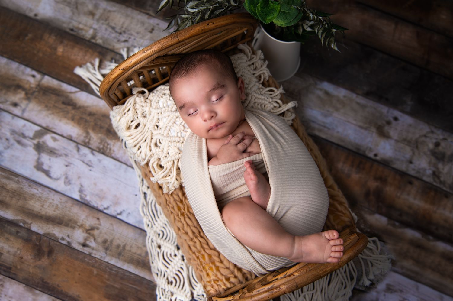 newborn photography showing baby wrapped in neutral fabric lying in rustic woven bed against wooden backdrop.