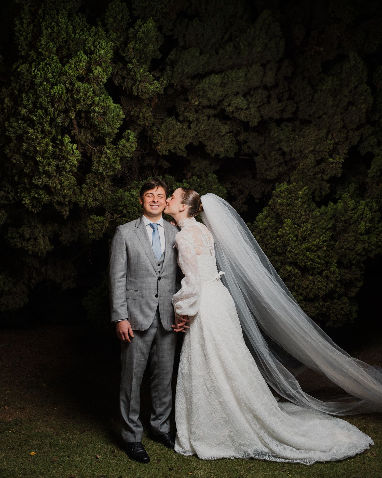 A couple in wedding attire pose together against a dark garden background with a flowing white veil and elegant dress.