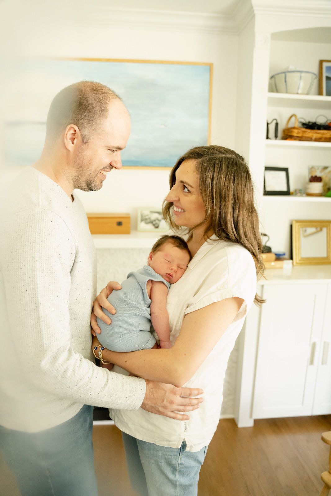 A tender family moment at home as parents share an intimate embrace while holding their sleeping newborn baby at lifestyle newborn session