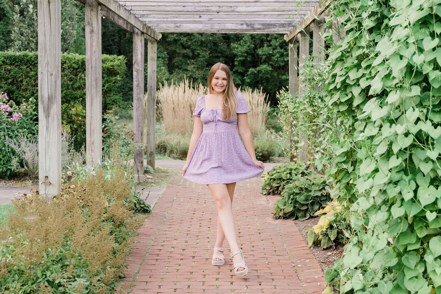 Lake High School senior in a flowing lavender dress twirls beneath a pergola covered in climbing vines along a brick pathway.