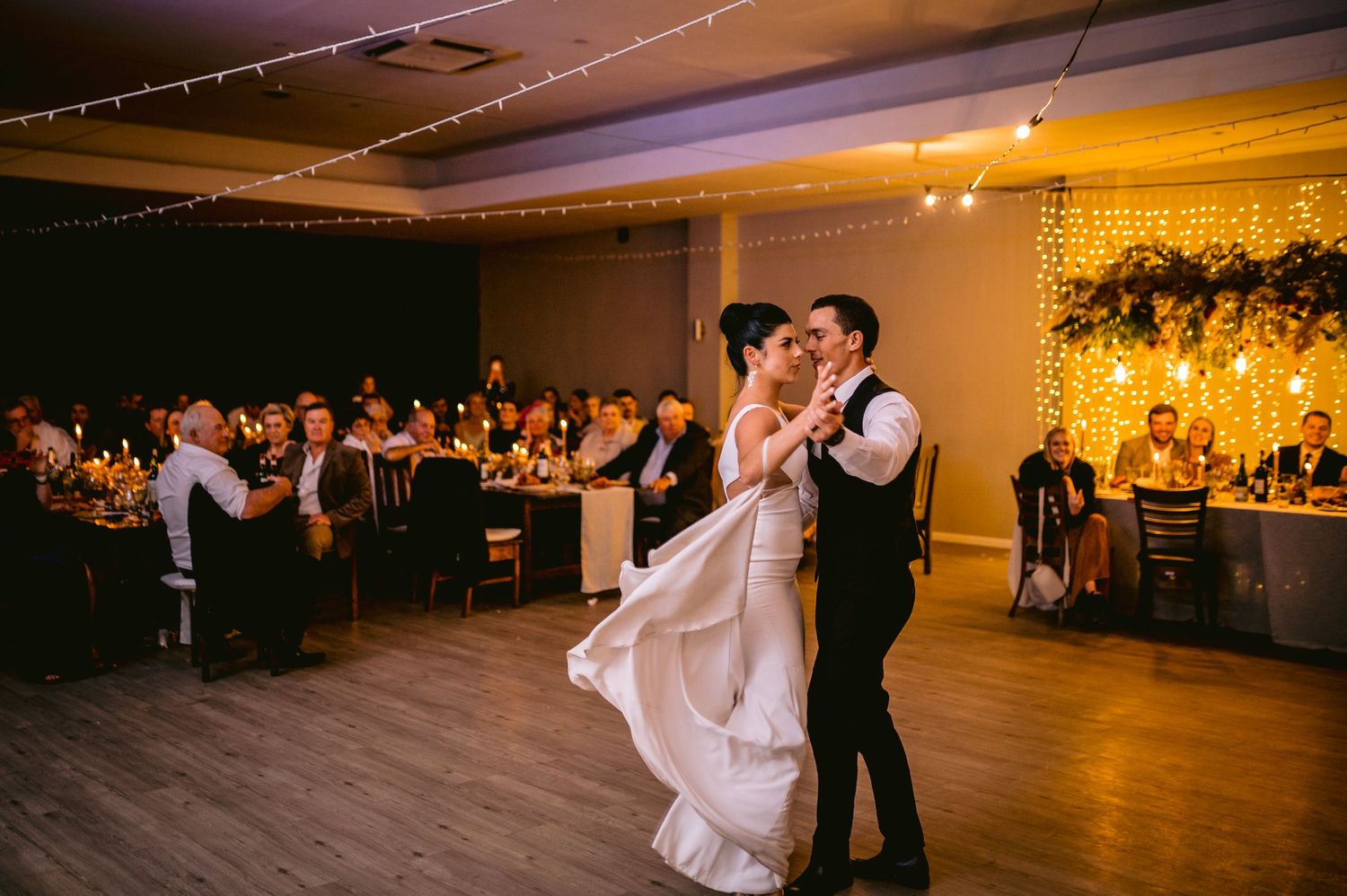 A couple shares their first dance at an elegant indoor wedding reception with warm lighting and floral decor.