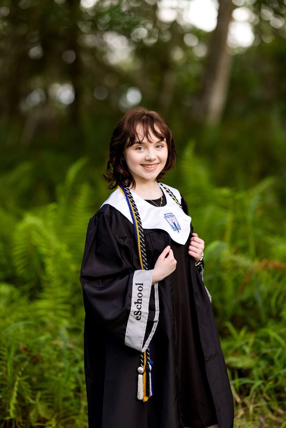 Graduate wearing ceremonial gown and cords stands among ferns in outdoor setting. Photos by Tampa Photographer DigitalMyst Photography