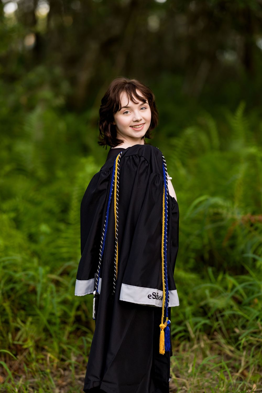 Graduate in black gown with honor cords poses against natural green backdrop. Photos by Tampa Photographer DigitalMyst Photography