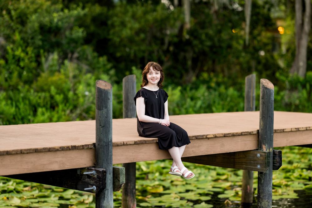 Student in graduation attire sits casually on edge of wooden dock over lily pads. Photos by Tampa Photographer DigitalMyst Photography