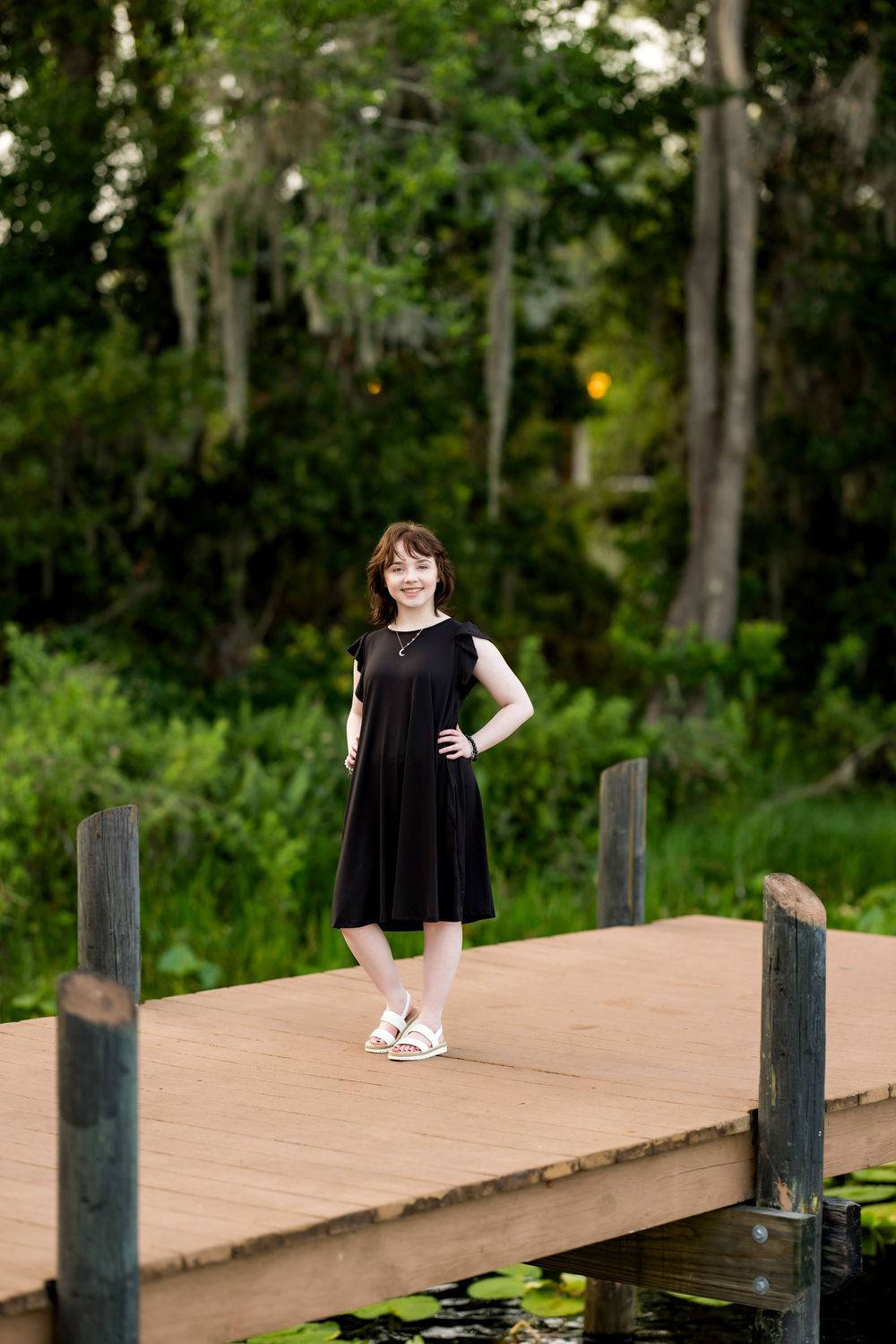 Graduate in a black dress stands on a wooden dock surrounded by lush greenery. Photos by Tampa Photographer DigitalMyst Photography