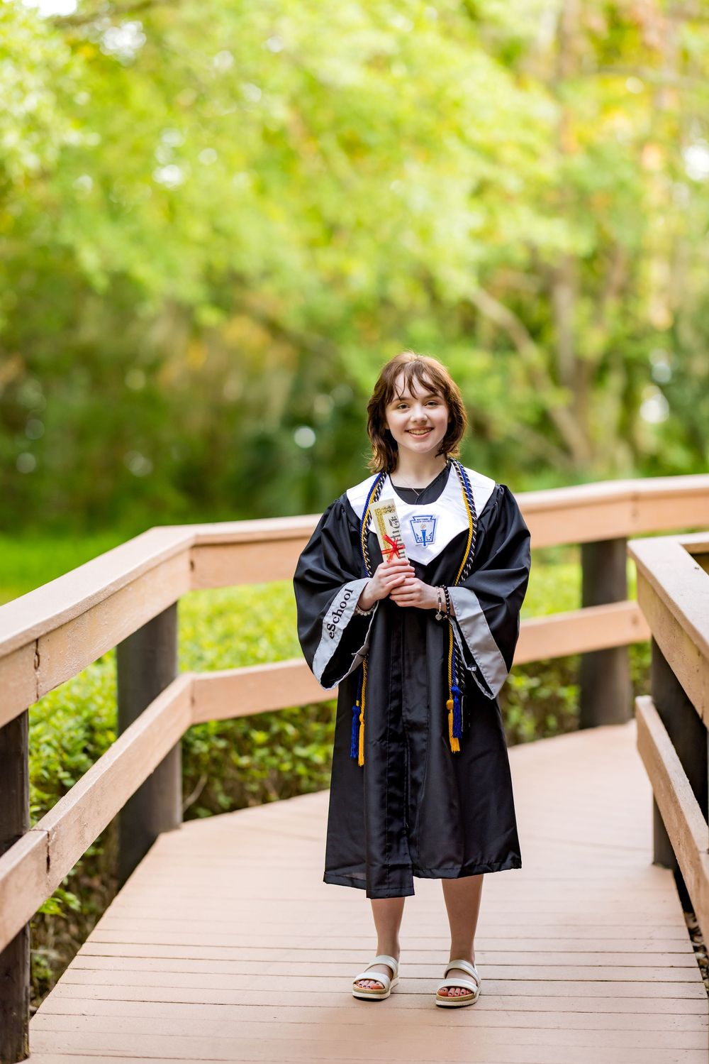 Graduate in black cap and gown stands smiling on a wooden bridge surrounded by lush greenery. Photos by Tampa's DigitalMyst Photography