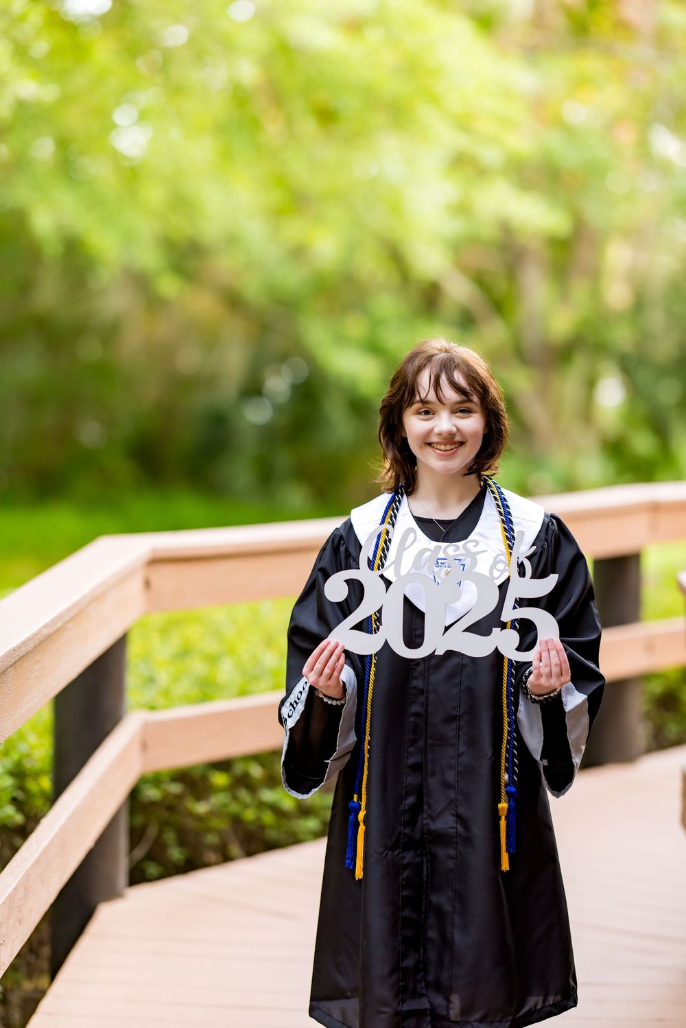Graduate wearing academic regalia holds up 2025 numbers while standing on outdoor bridge. Photos by Tampa DigitalMyst Photography